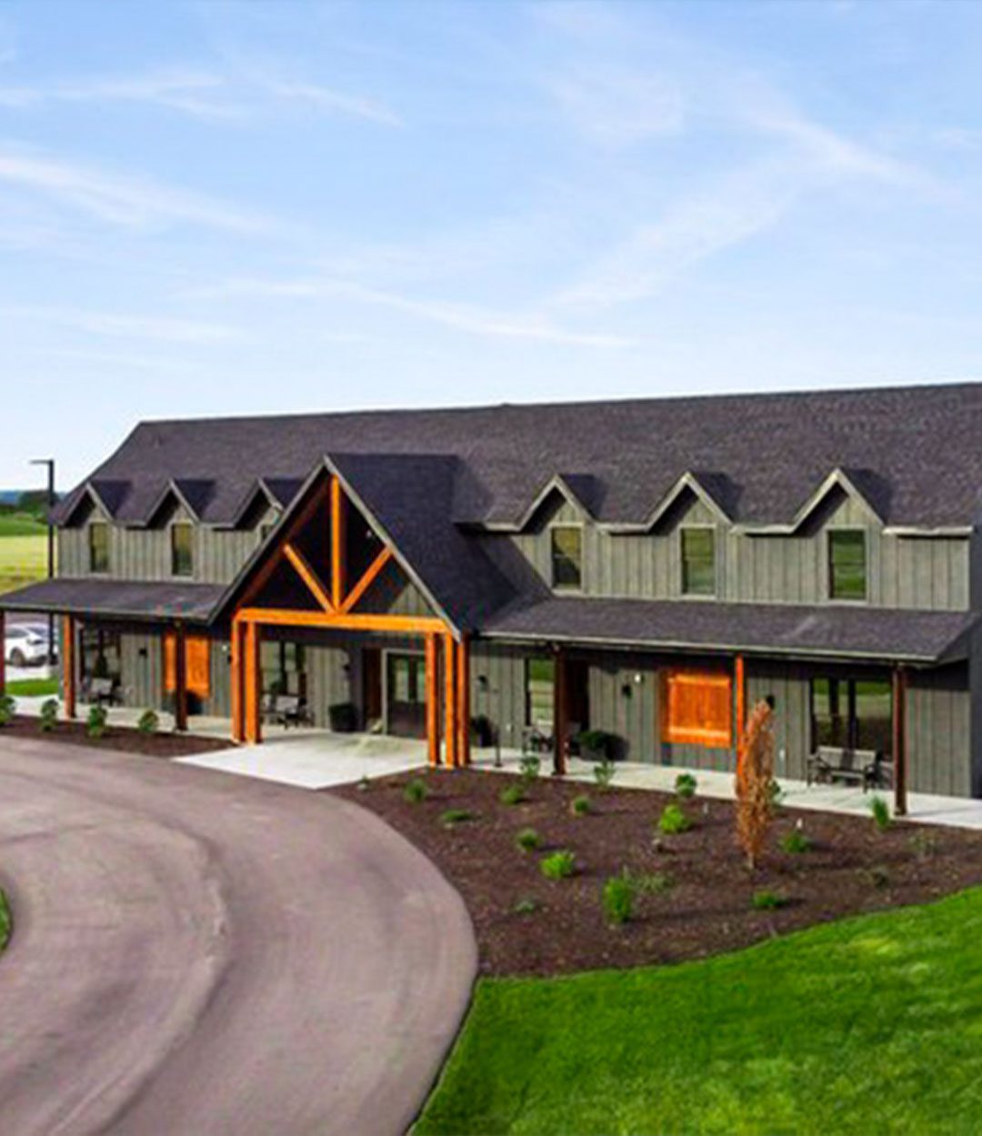 Rustic modern event venue with wood accents, dormer windows, and landscaped circular driveway surrounded by lush green fields.