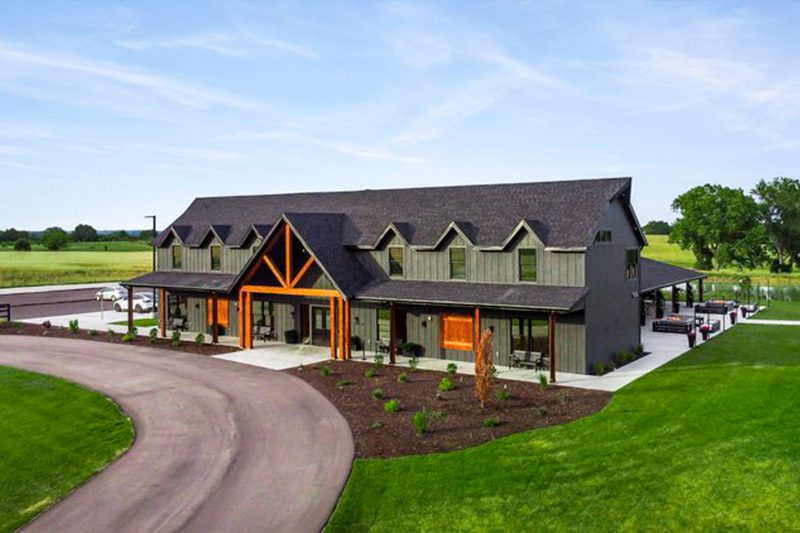 Aerial view of Haven Event Venue, a modern rustic building with wood accents and large windows, surrounded by landscaped greenery and open fields under a clear sky.