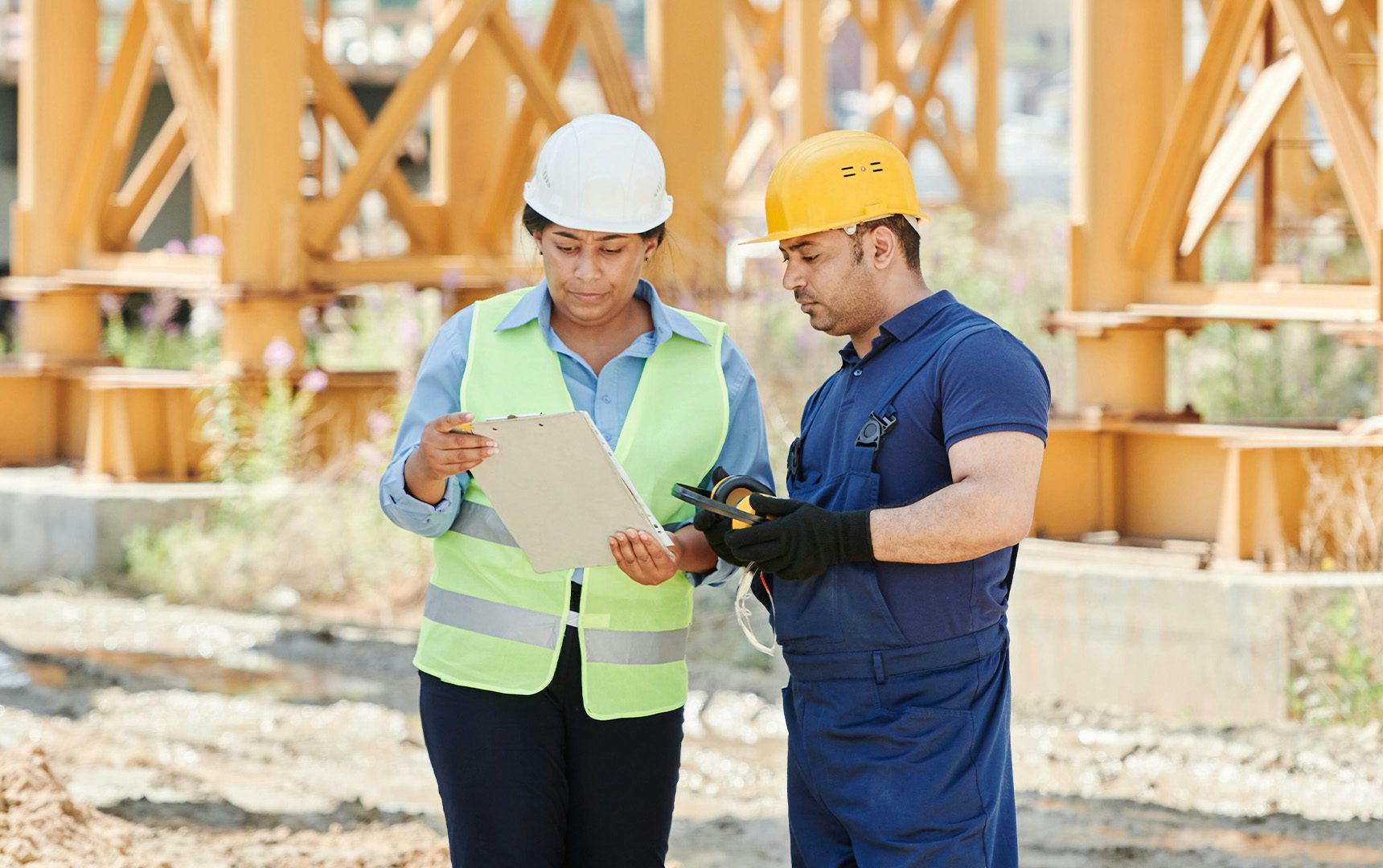 Site Supervision Two construction workers wearing safety gear reviewing a clipboard and measuring tool on a jobsite, with structural steel beams in the background.