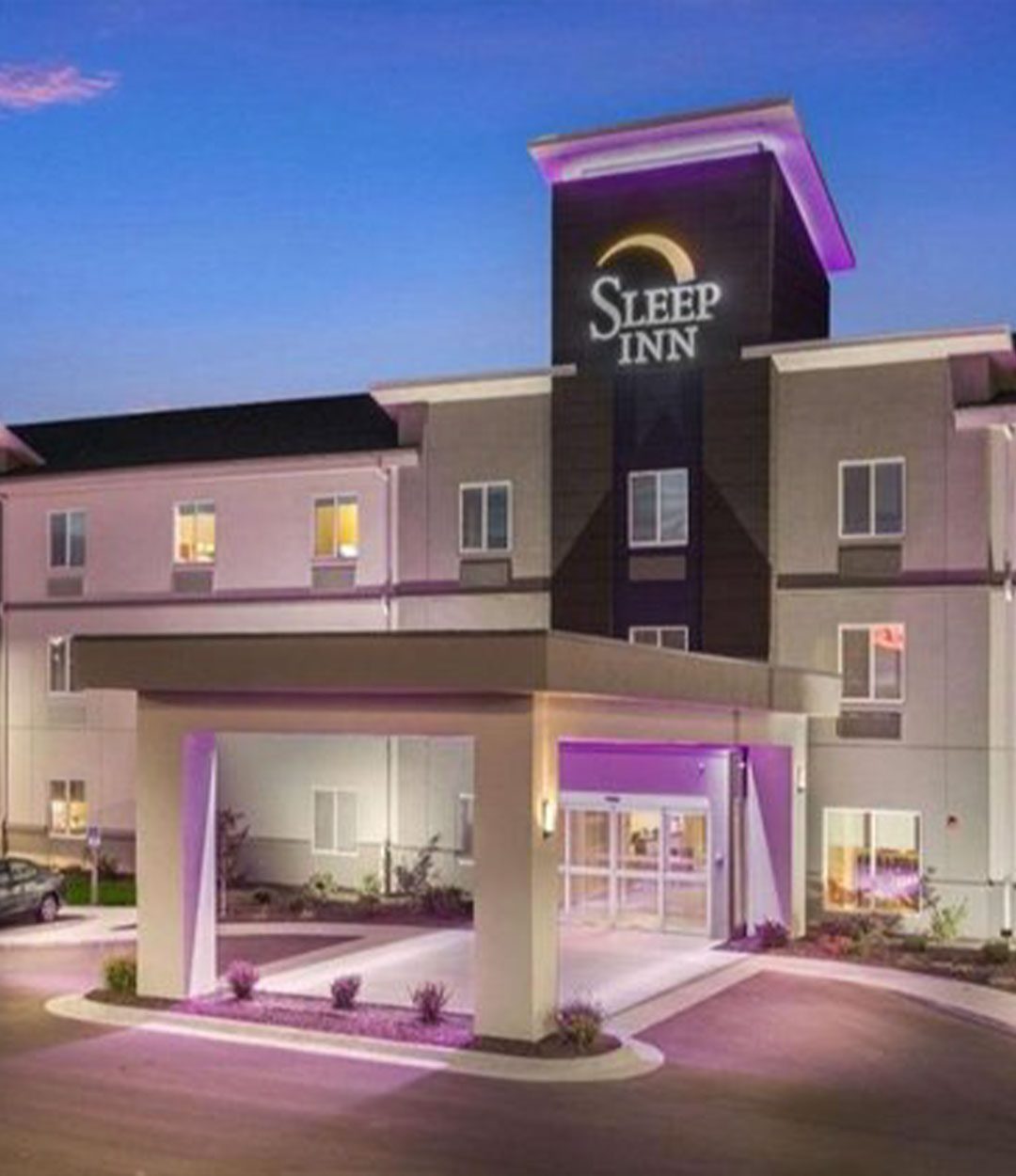 Modern Sleep Inn hotel exterior at dusk, illuminated with purple accent lighting and a large front canopy entrance under a twilight sky.