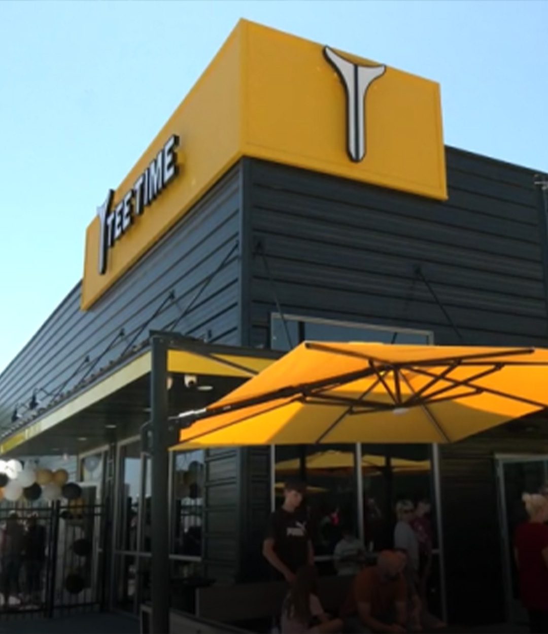 Modern black building with yellow signage for "Tee Time," featuring a crowd gathered under matching yellow umbrellas and balloons decorating the entrance.
