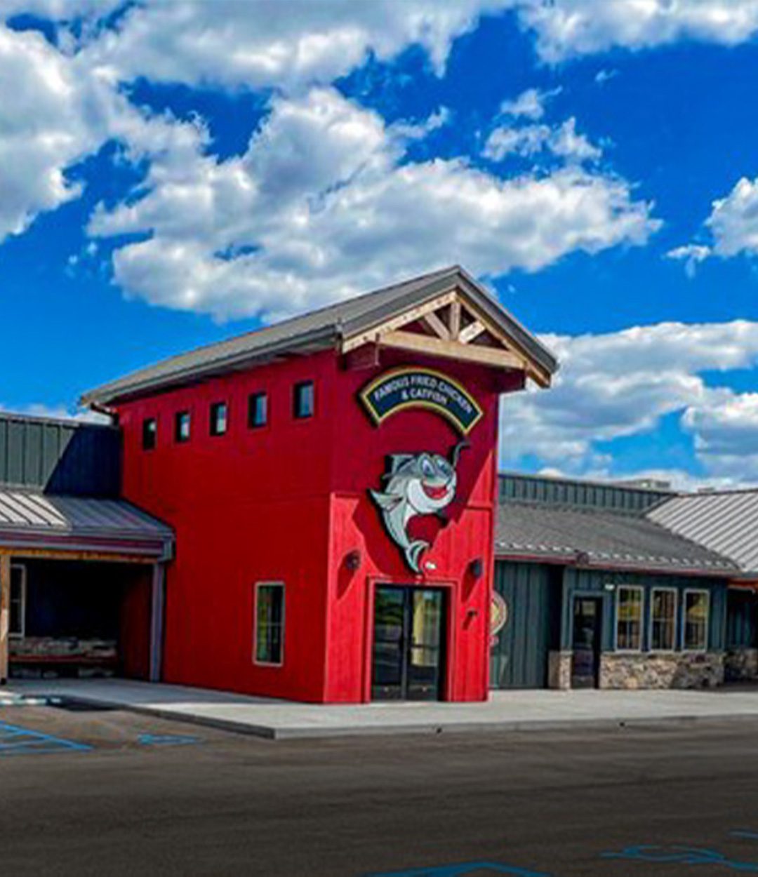 Bright red entry tower with cartoon fish logo and sign reading "Famous Fried Chicken & Catfish," attached to a rustic-style building under a blue sky with clouds.