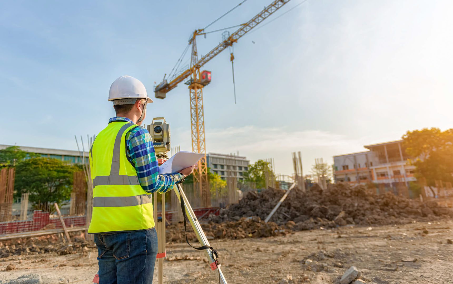 Value Engineering Construction site engineer using surveying equipment and reviewing plans, with a tower crane and building framework visible in the background under a bright sky.