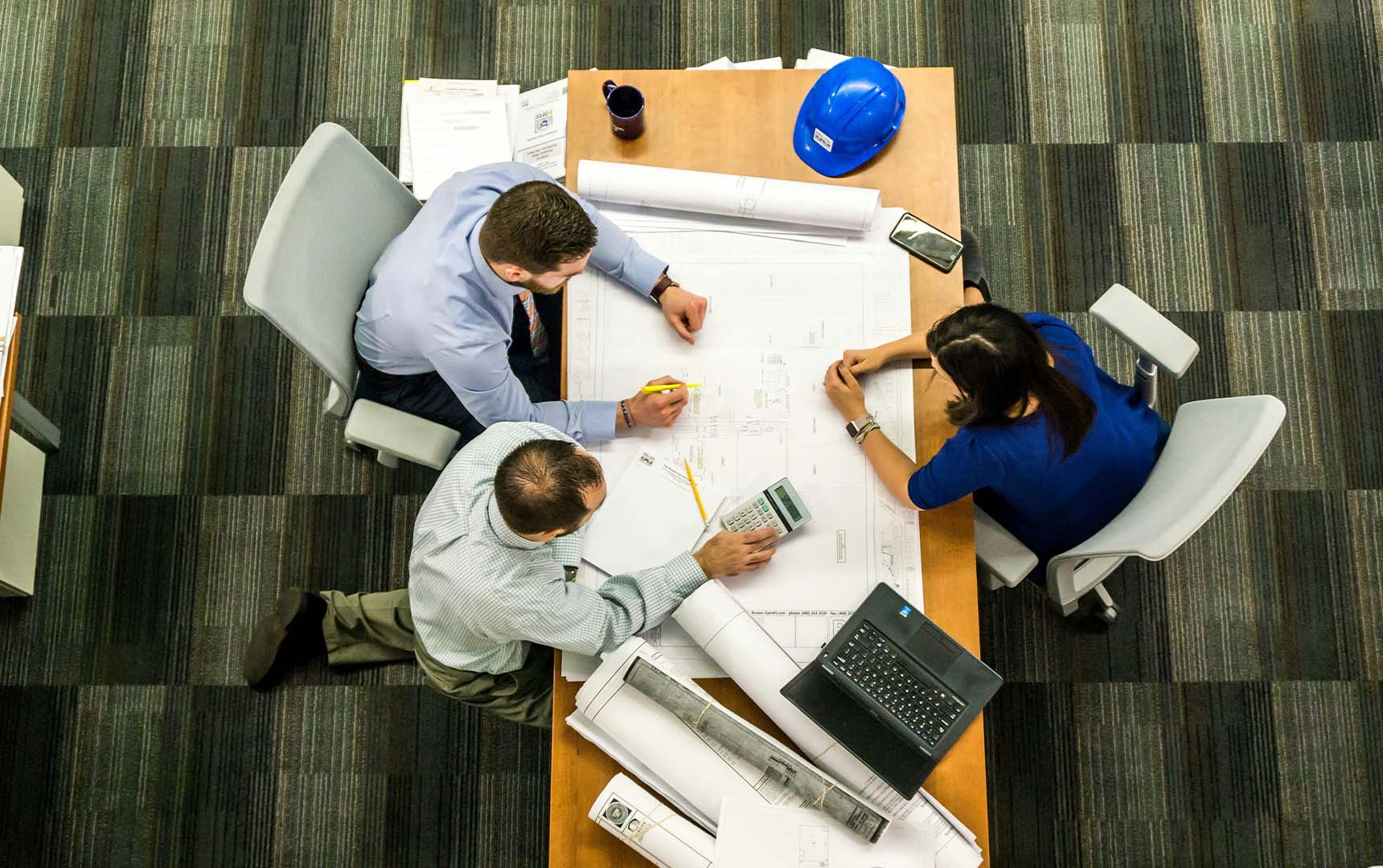 Value Engineering Three professionals gathered around a table reviewing blueprints and using a calculator during a construction planning meeting, with a blue hard hat and laptop nearby.